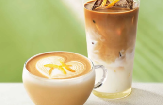 Hot latte in a glass cup with latte art beside a tall iced coffee with milk on a light surface and a green background
