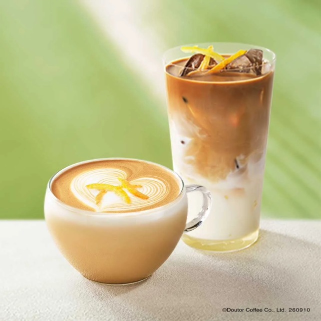 Hot latte in a glass cup with latte art beside a tall iced coffee with milk on a light surface and a green background