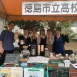 Six students in blue aprons and uniforms stand behind a coffee stall under an orange canopy smiling and posing with peace signs