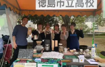 Six students in blue aprons and uniforms stand behind a coffee stall under an orange canopy, smiling and posing with peace signs.