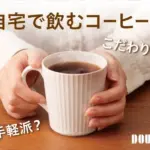 Hands hold a white mug of coffee on a wooden table with Japanese text asking if home brewed coffee should be artisanal or quick DOUTOR logo