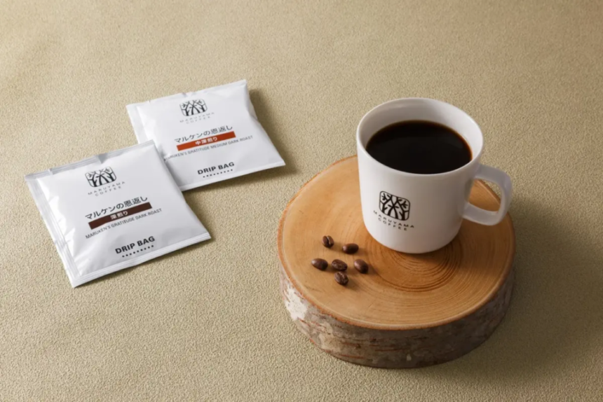 White mug of black coffee on a round wooden coaster with a few coffee beans and two drip bag coffee packets nearby