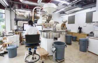 Person seated at a control station monitoring a large industrial coffee roaster in a bright workshop