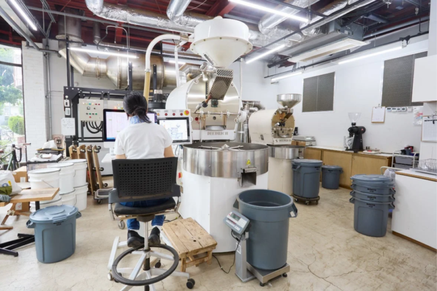 Person seated at a control station monitoring a large industrial coffee roaster in a bright workshop