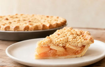 Slice of apple crumble pie on a white plate with a whole pie in the background