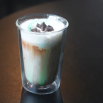 Tall glass with a layered milk drinkwhite foam on top brown and green layers topped with chocolate shavings on a dark table