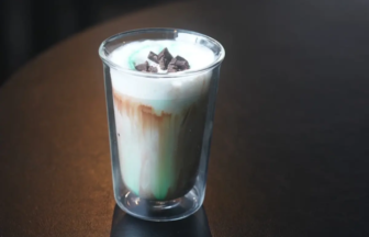 Tall glass with a layered milk drinkwhite foam on top brown and green layers topped with chocolate shavings on a dark table