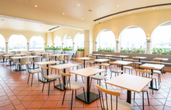Bright airy dining area with many square tables and padded chairs arched windows and potted plants along a long wall