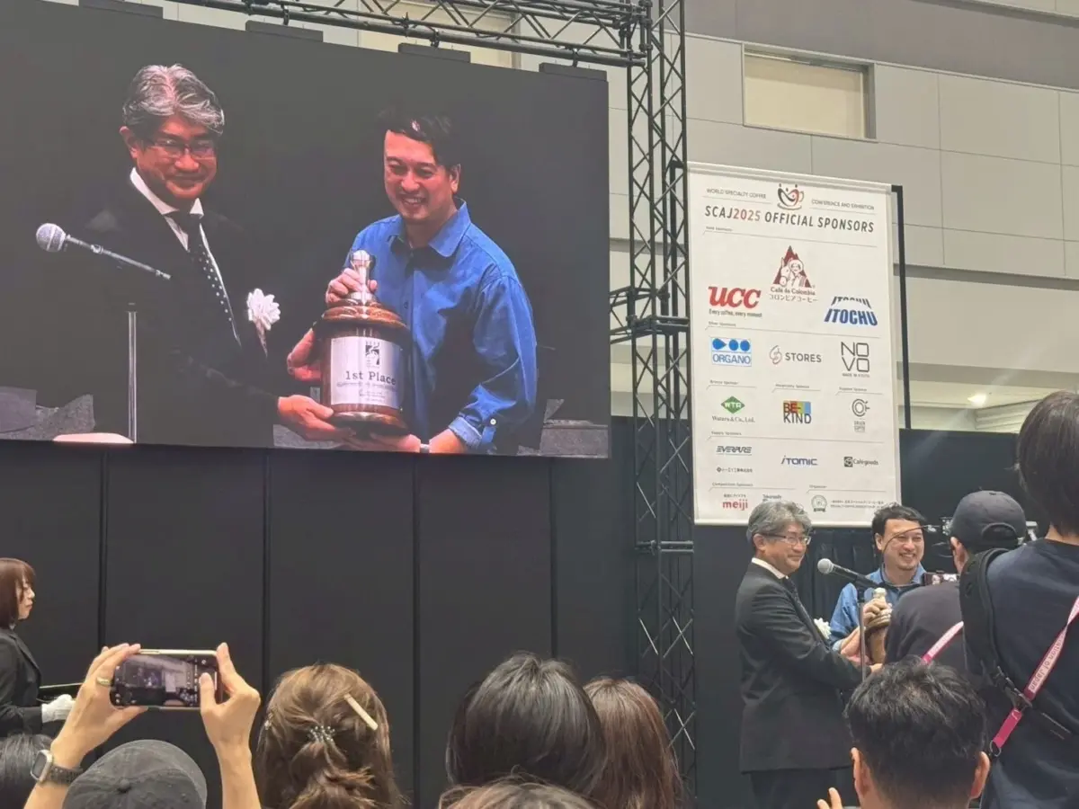 SCAJ2025のコーヒーイベント会場で、男性が「1st Place」のトロフィーを受け取っている表彰式の様子。
