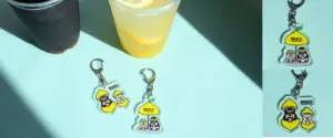 Two drinks on a turquoise surface one dark one yellow lemonade with three Becks Coffee Shop keychains featuring lemon shapes and cartoon characters nearby