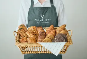 Person in a gray apron holding a wicker basket filled with assorted bread loaves and a white cloth lining the basket