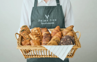 Person in a dark apron with the Prime Five Tullys Coffee logo holds a wicker basket filled with assorted breads including croissants and baguettes