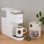 White Keurig style single serve coffee maker on a wooden counter with a mug labeled Tullys coffee under the dispenser plus a Tullys pod box nearby and a plant in the background
