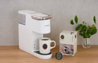 White Keurig style single serve coffee maker on a wooden counter with a mug labeled Tullys coffee under the dispenser plus a Tullys pod box nearby and a plant in the background