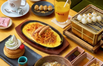 A wooden table filled with Japanese cafe dishes: omurice with noodles, a coffee cup, orange juice, dango skewers, and a dessert with cream crown; green couch in background.