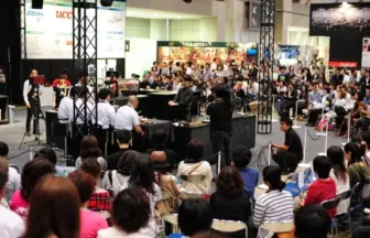Crowd watches a tabletop competition in a convention hall; players at tables, judges, camera crew, with sponsor banners in the background.