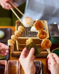 Skewers of grilled dango with a sweet glaze one wrapped in seaweed held over a small grill in the background