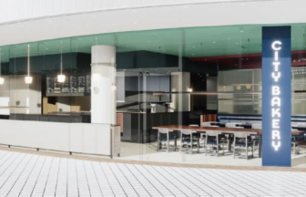 Modern cafe interior with a curved glass storefront white countertops menu boards and a row of high tables along a blue vertical sign that reads CITY BAKERY on the right side
