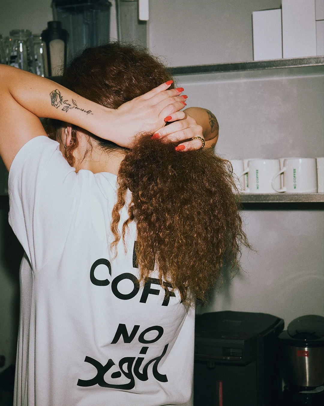 「NO COFFEE NO X-girl」Tシャツを着用し髪を結ぶ女性の後ろ姿