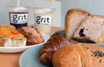 Brunch spread with a croissant sesame bun blueberry loaf and veggie topped toast on plates plus two cups labeled grit coffee in the background