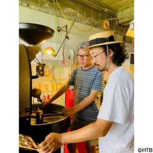 Two men operate a coffee roasting machine in a workshop adjusting its controls