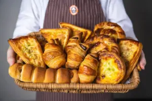 Baker in a striped apron presents a wicker basket full of croissants and cheese filled pastries