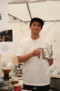 Man in a white t shirt and black cap posing with a metal gooseneck kettle at a coffee booth with pouring over gear on the table in front of him