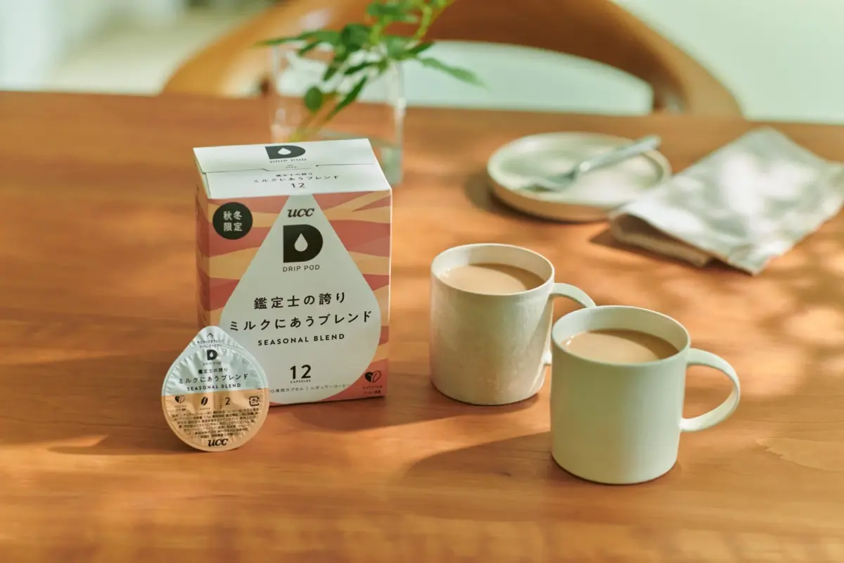 木のテーブルに置かれたUCC DRIP PODの「ミルクにあうブレンド」コーヒーの箱とカプセル、そしてミルク入りのコーヒーが入ったマグカップが2つ写っています。