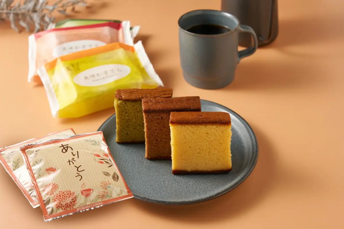 プレーン、抹茶、チョコレート味の3種のカステラが皿に並び、個包装のカステラや「ありがとう」と書かれたコーヒー小袋、温かいコーヒーが添えられた、和やかなティータイムの様子