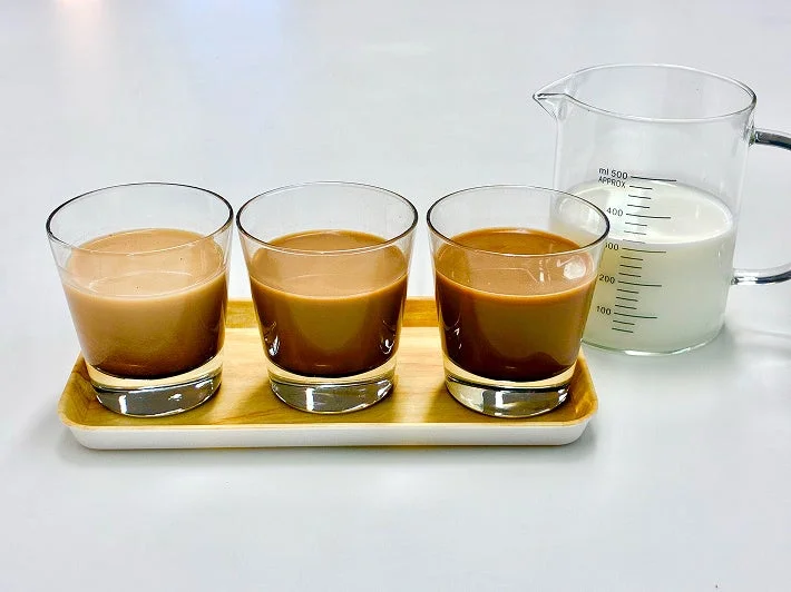 木のトレイに異なる濃さのカフェオレのような飲み物が3つのグラスに入っており、隣にはミルクが入った計量カップが置かれています。