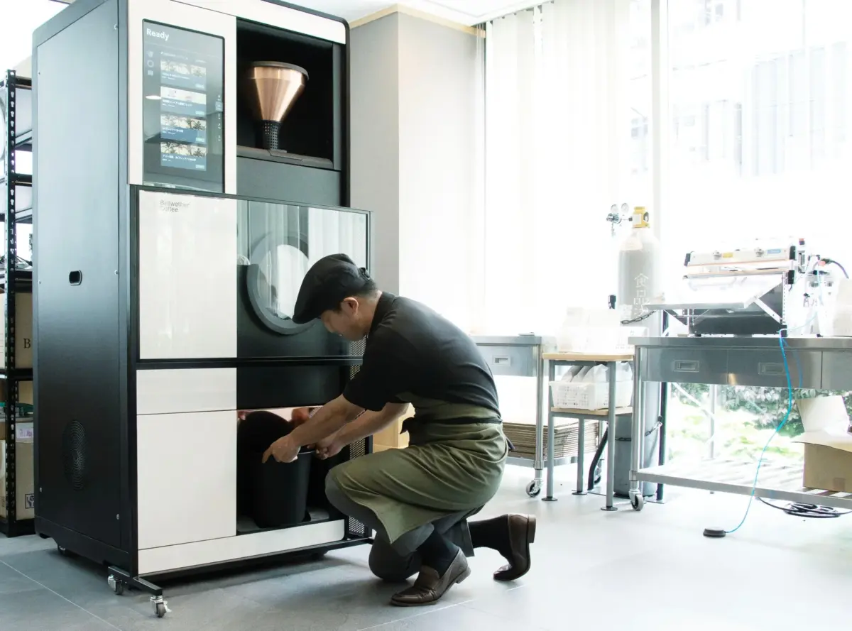 明るい作業スペースで、男性がエプロンと帽子を着用し、Bellwether Coffeeの大型コーヒー焙煎機を操作している様子。機械にコーヒー豆を投入していると見られます。