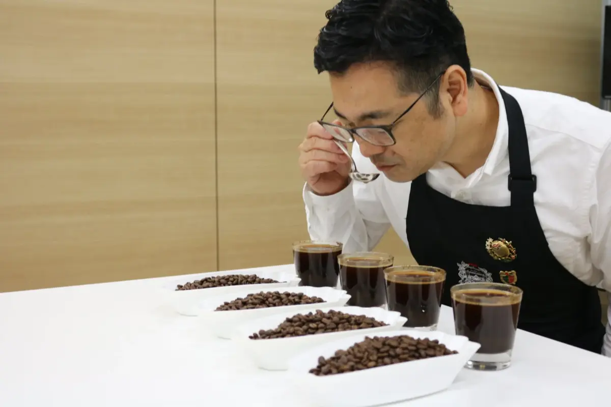 男性がコーヒーのカッピングを行い、スプーンでコーヒーの香りを確かめている様子です。複数のコーヒー豆と抽出されたコーヒーが並べられ、品質評価をしている専門的なシーンがうかがえます。