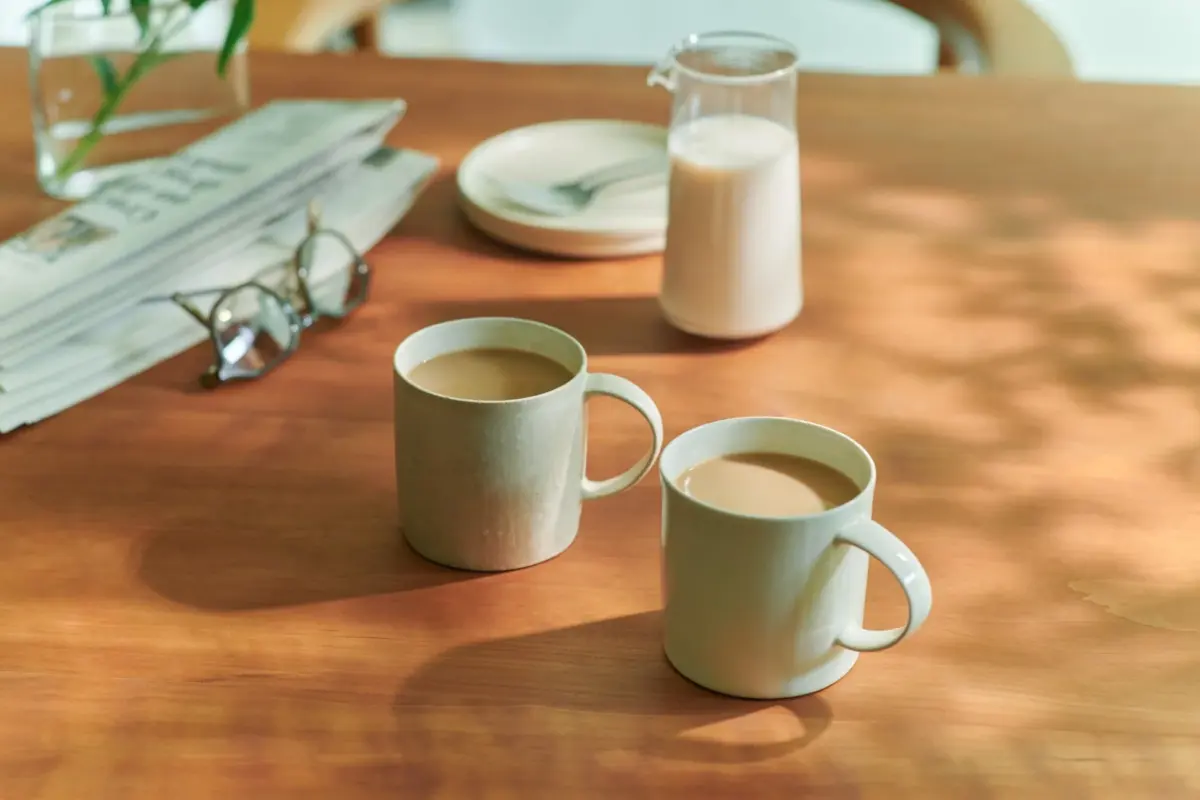 木のテーブルに置かれた2つのコーヒーカップ、新聞、眼鏡、ミルクが写っています。